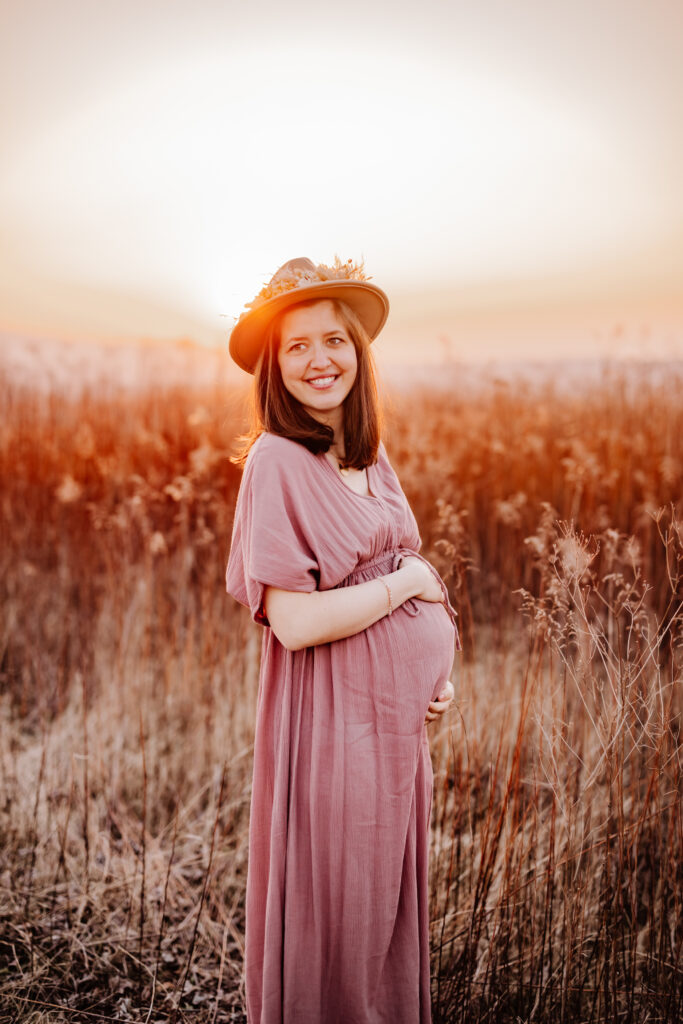 Sarah Glück schwanger in einem Feld bei Sonnenuntergang, die Hände am Babybauch